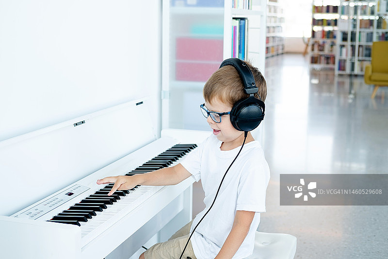 戴眼镜、大耳机、穿白色T恤的小男孩在音乐课上弹奏白色数码钢琴图片素材