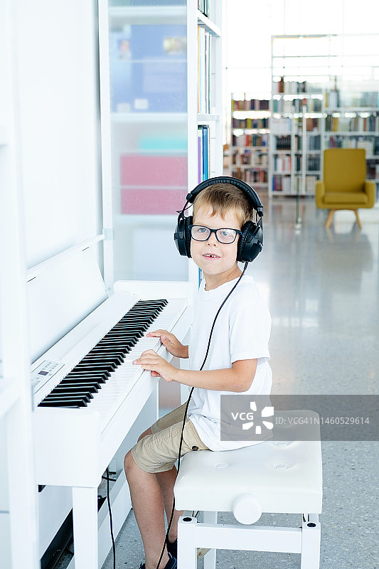 戴眼镜、大耳机、穿白色T恤的小男孩在音乐课上弹奏白色数码钢琴图片素材