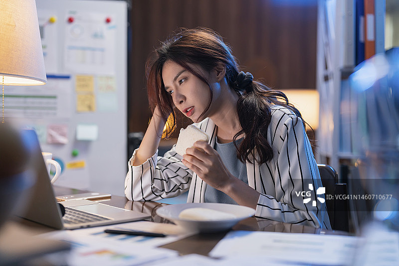 年轻的亚洲成年女性深夜在家工作，忙碌地吃三明治图片素材
