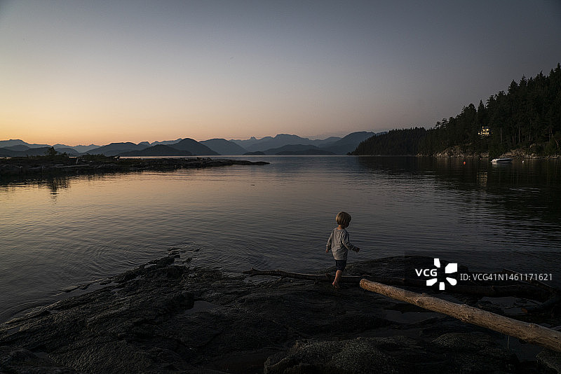 黄昏时分，男孩在水边岩石上行走，背景是群山图片素材