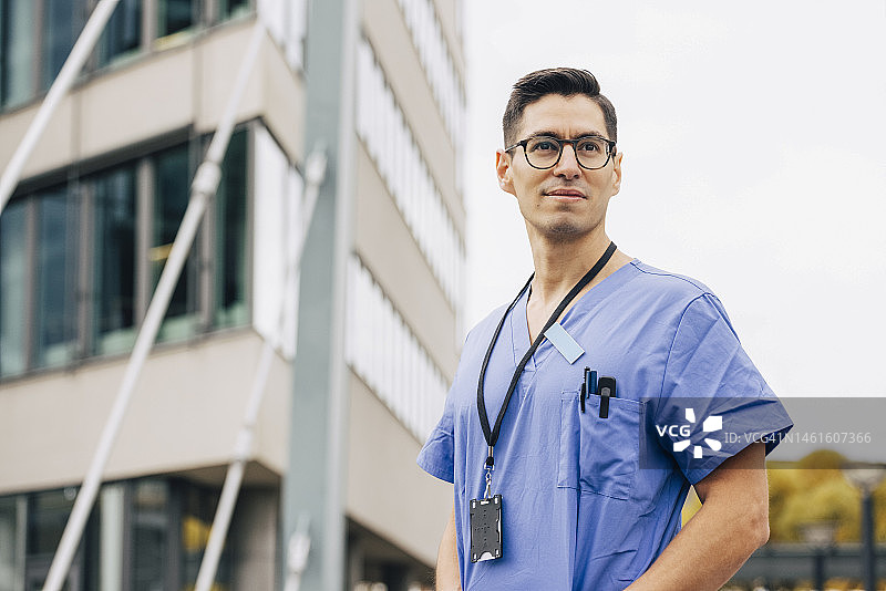 自信的男医生穿着蓝色工作服，在医院大楼前看向远方图片素材