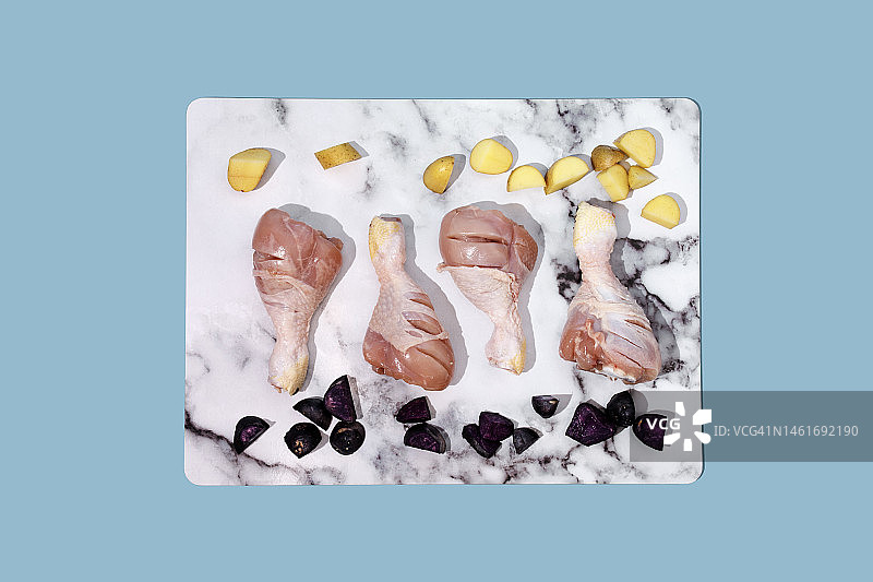 蓝色背景上的生鸡腿、土豆和紫土豆——食物摆拍图片素材