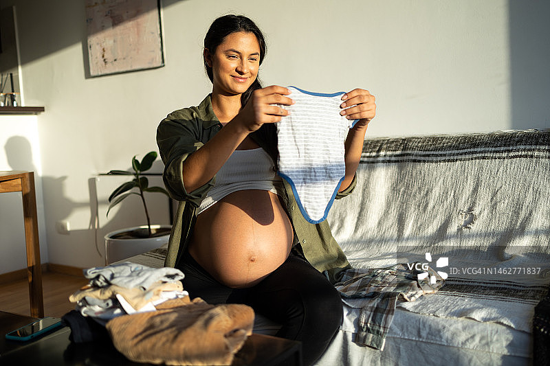 年轻孕妇坐在床上叠衣服，为宝宝准备和挑选小衣服，布置婴儿房图片素材