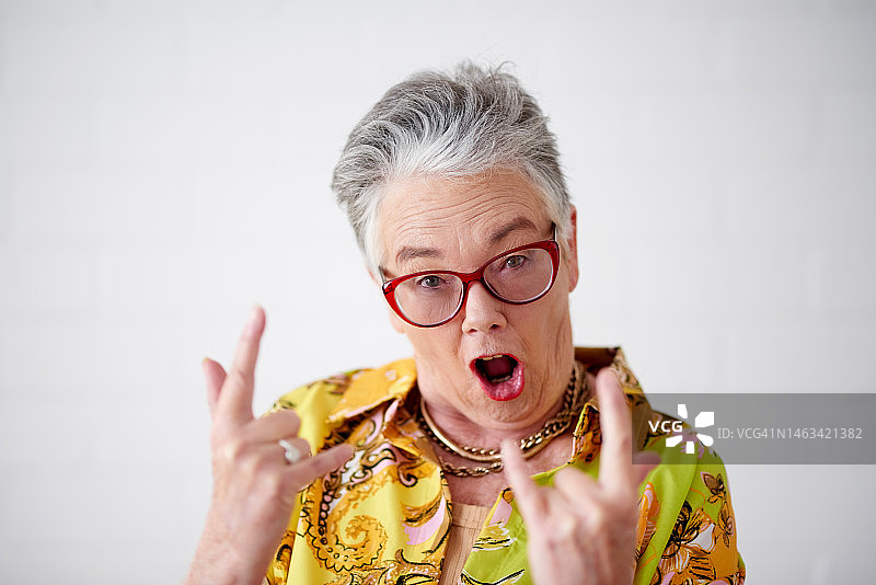 摇滚手势，年长的白人女性在影棚中展现金属、朋克和疯狂的风格。孤立的白色背景和一个兴奋的老年人表情符号，表达酷和准备庆祝的感觉图片素材