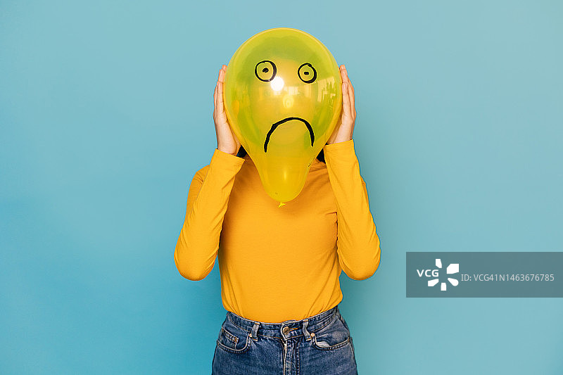 年轻女子躲在画着悲伤面孔的气球后面。负面情绪概念图片素材