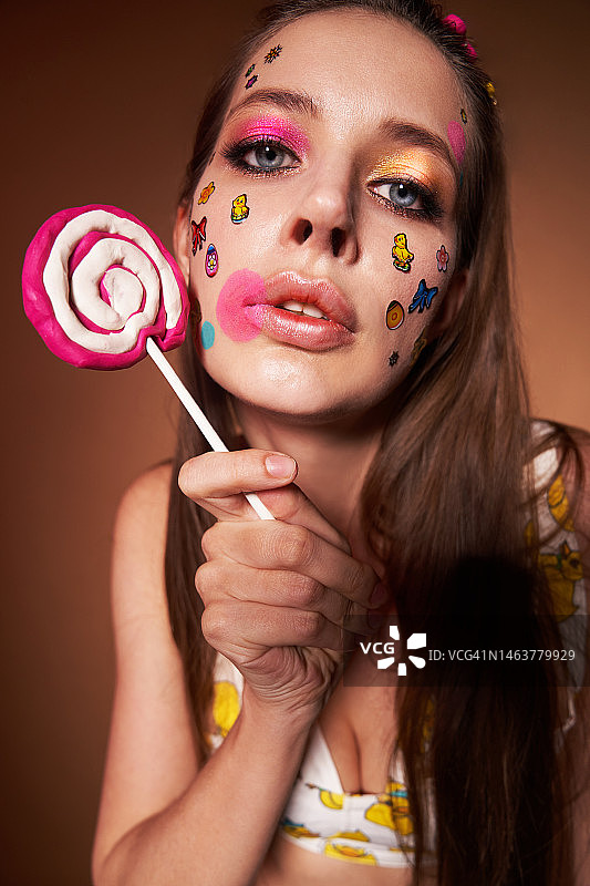 浓妆艳抹的年轻女子，脸上贴着贴纸，拿着一个大甜糖果，穿着一件鲜艳的上衣。甜蜜生活，青春的概念，具有复制空间的时尚历史。图片素材