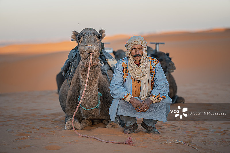 摩洛哥骆驼司机蹲在撒哈拉沙漠，骆驼看着镜头图片素材