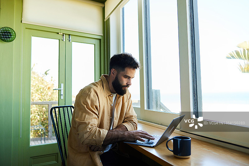 年轻男人在家中窗边的桌旁使用笔记本电脑图片素材