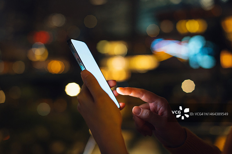 夜晚，市中心街区，女子用智能手机浏览屏幕，城市建筑的散景灯光背景。生活方式与科技图片素材