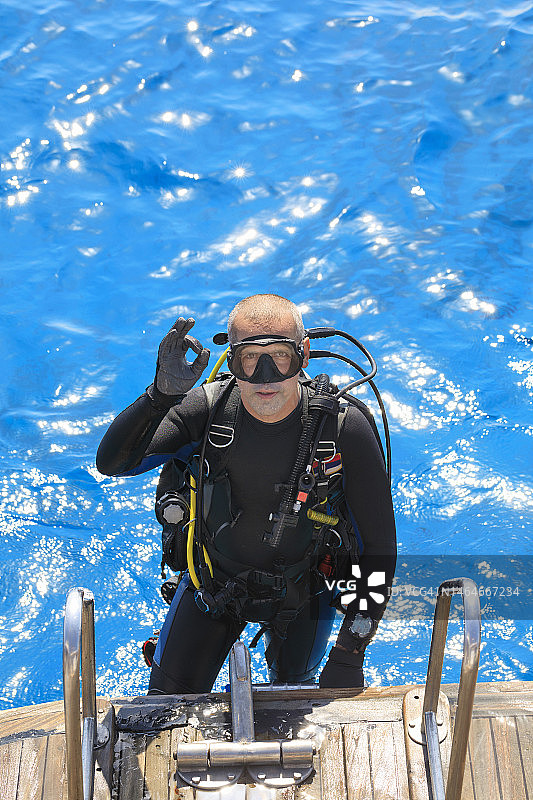 准备水肺潜水的中年英俊男运动员潜水员，显示OK手势。美丽的热带蓝色海洋，珊瑚礁在背景中。水上运动图片素材