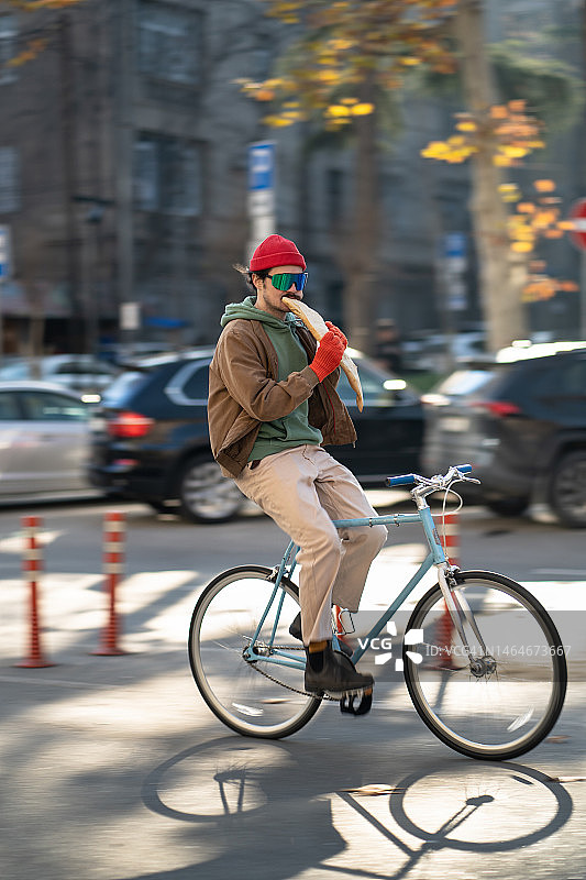 千禧一代时髦的城市自行车手在清晨的城市骑自行车时吃新鲜出炉的面包图片素材