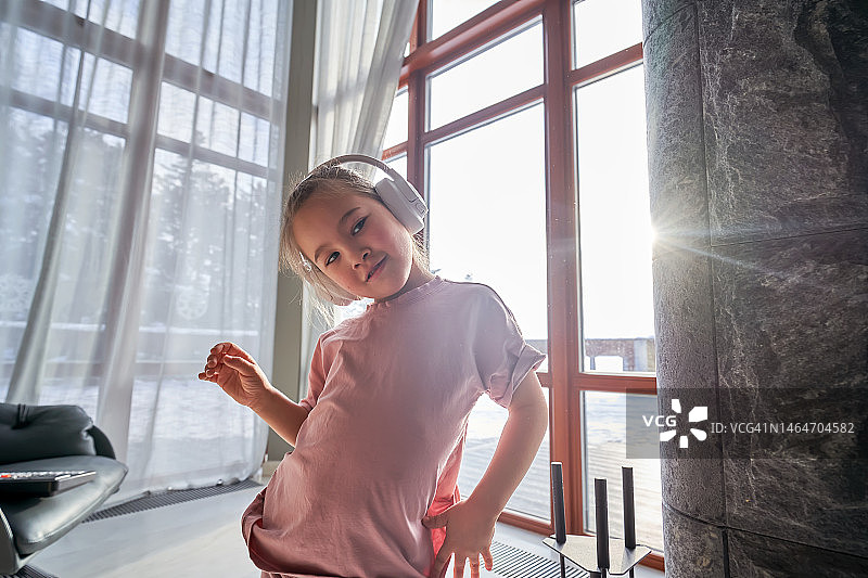 一个穿着粉红色西装的6岁女孩站在全景窗户的背景前，戴着白色大号头戴式耳机听音乐图片素材