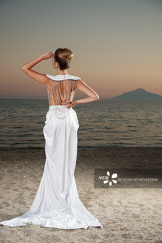 穿白色长裙、戴珍珠首饰的年轻女子在黄昏时分站在沙滩上的背影图片素材