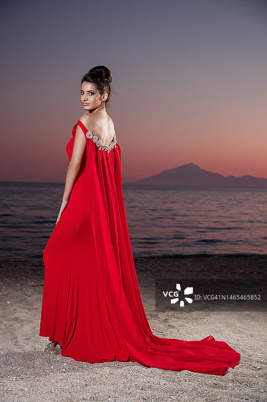 穿着红色无袖长裙的年轻漂亮女子站在黄昏的沙滩上，回头看图片素材