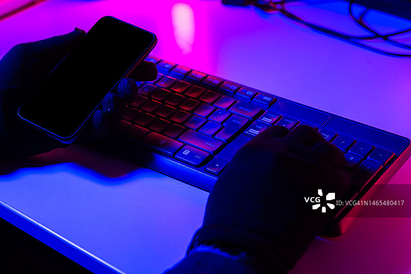 戴着手套的手拿着手机，在电脑键盘上打字并窃取信息，蓝粉色灯光照亮。网络安全、盗窃、黑客、身份和犯罪概念。图片素材