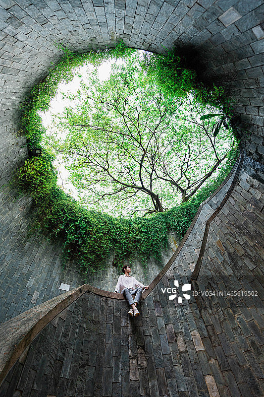 年轻亚洲男子坐在螺旋楼梯的巨树下。图片素材