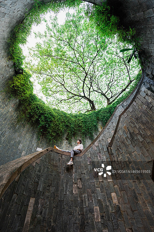 年轻亚洲男子坐在螺旋楼梯的巨树下。图片素材