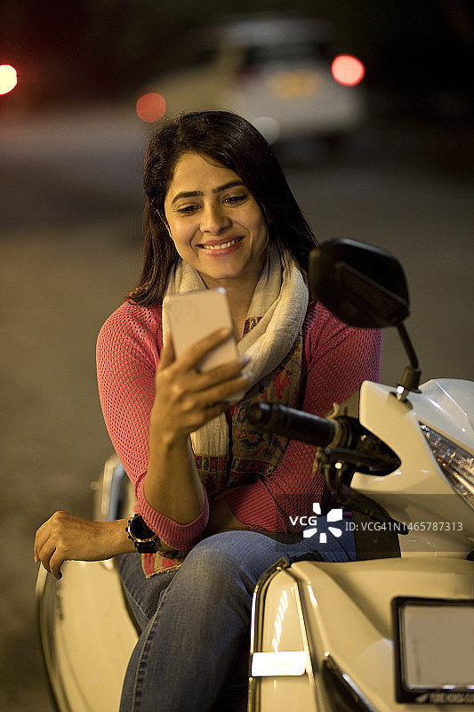 晚上坐在摩托车上用手机聊天的快乐女人图片素材