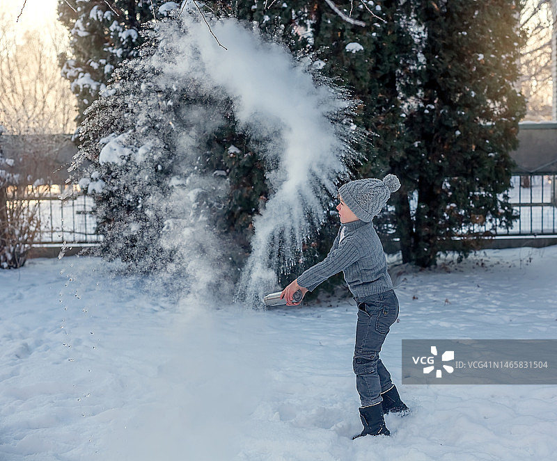 在阳光明媚的冬日，穿着冬季针织服装的男孩在院子里圣诞树旁，从保温瓶中倒出开水图片素材