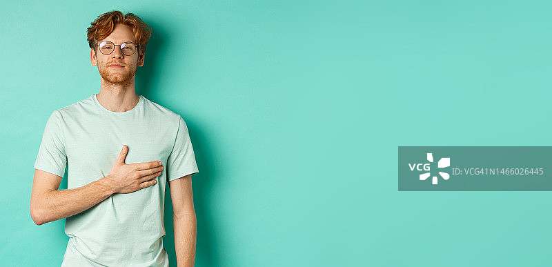 自豪的年轻红发男子戴着眼镜，手放在胸前，正在倾听图片素材