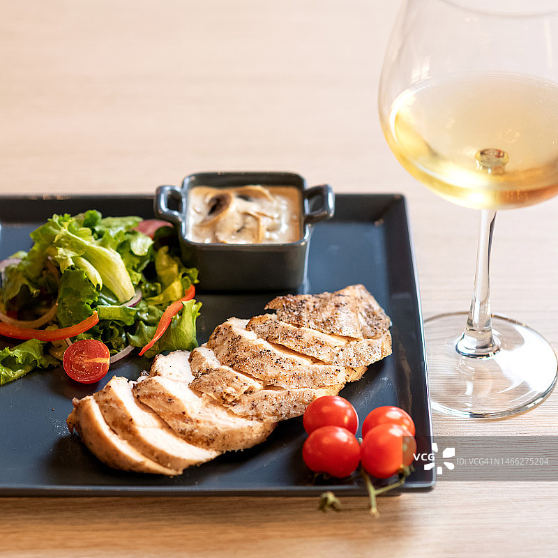 餐厅晚餐：烤鸡胸肉、酱汁和新鲜蔬菜沙拉。黑色方盘盛放的菜肴和木桌上的白葡萄酒杯。健康食品，侧视图图片素材