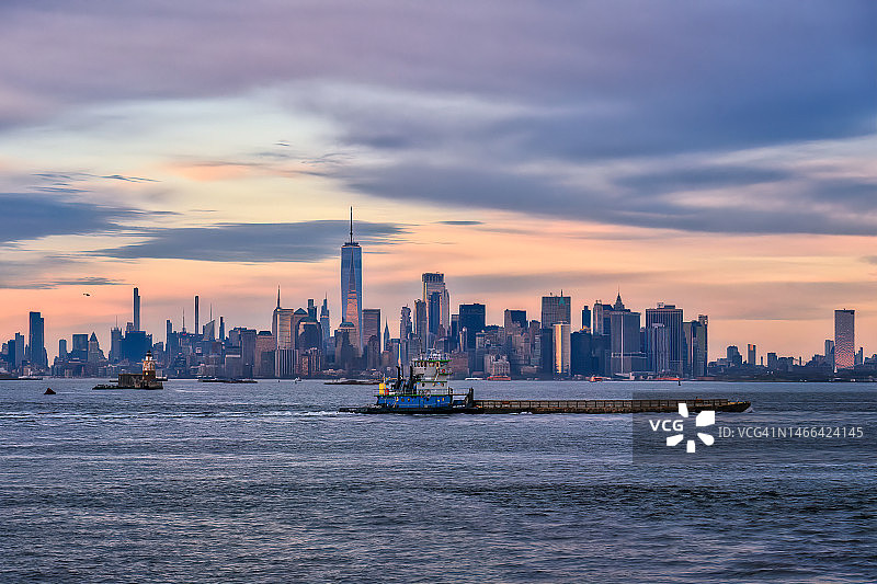 夕阳下的纽约港，拖船正在推送驳船，远处是曼哈顿下城图片素材