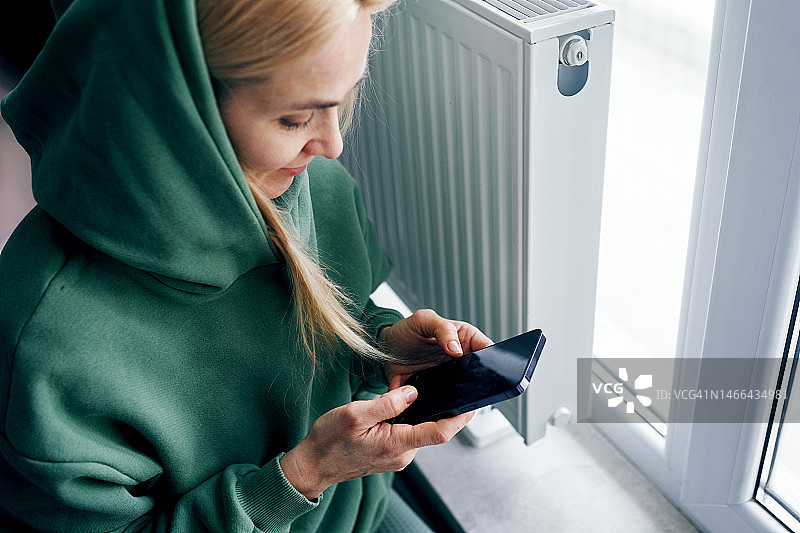冬天在家，在暖气片旁使用手机的年轻女子肖像图片素材