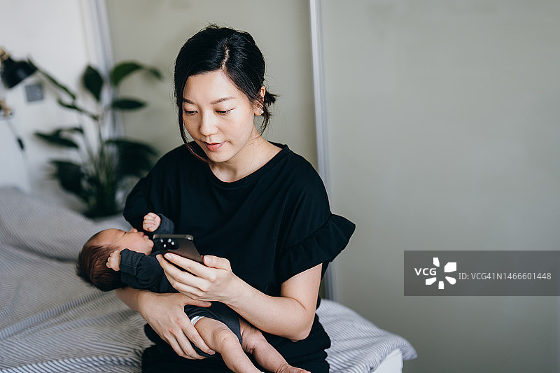 年轻的亚洲母亲在卧室里用智能手机，同时抱着正在睡觉的新生女婴。母爱，科技家庭生活方式图片素材