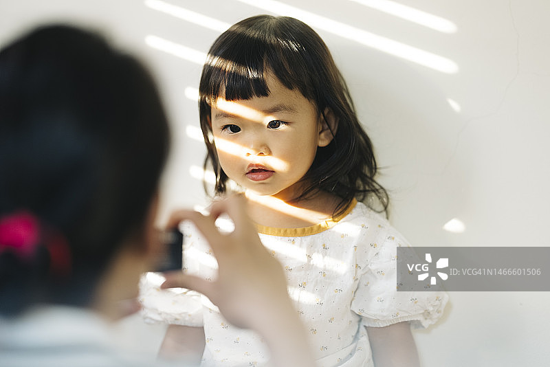 积极乐观的情绪：快乐地在家里的起居室使用小型玩具相机的亚洲女童，享受家庭生活，与母亲共度美好时光图片素材