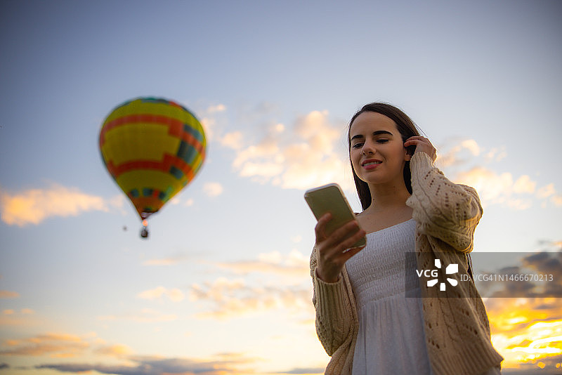 日出时分，年轻女子使用手机，背景是飞行的巨大热气球图片素材