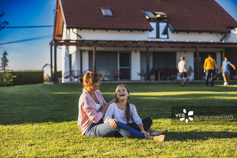 母亲在后院的草坪上为女儿编辫子，父亲在露台前与两个儿子踢足球图片素材