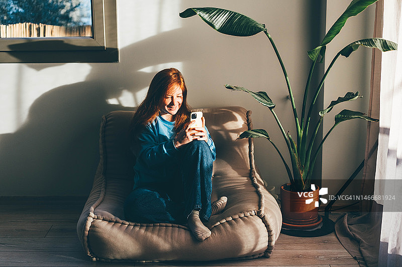 快乐的年轻女子坐在沙发上，在家使用智能手机图片素材