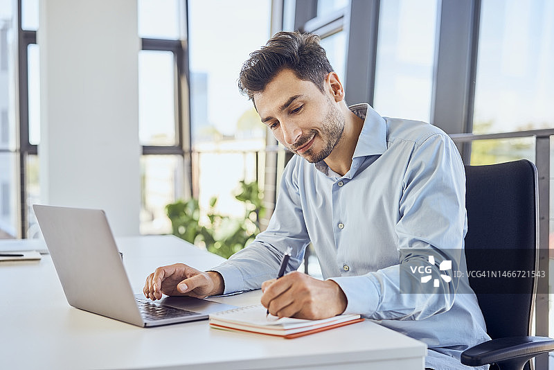 商人在办公室里用笔记本电脑在日记本上记笔记图片素材