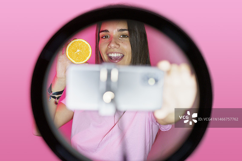 微笑的网红在环形灯下用智能手机拍摄橙子图片素材