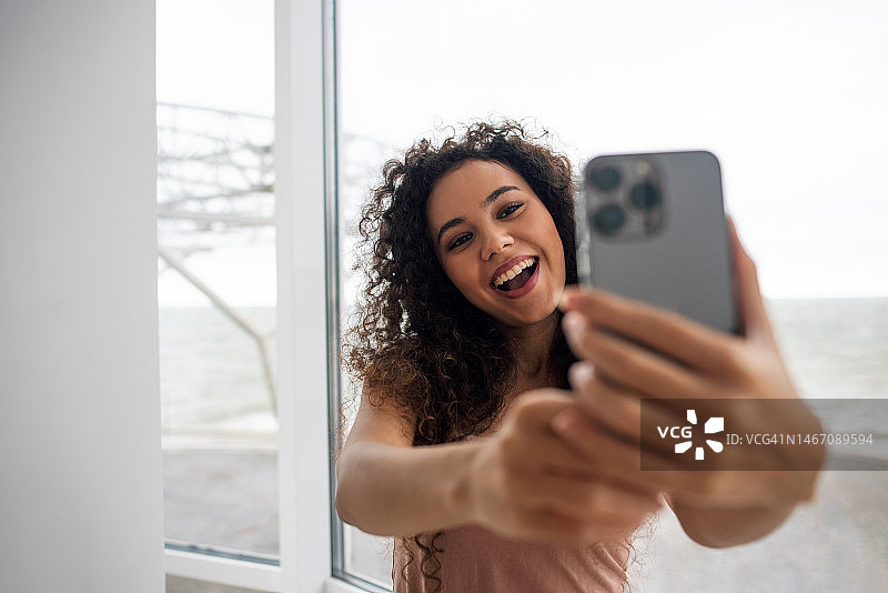 年轻黑人女性在窗边自拍，使用智能手机进行社交和视频沟通图片素材