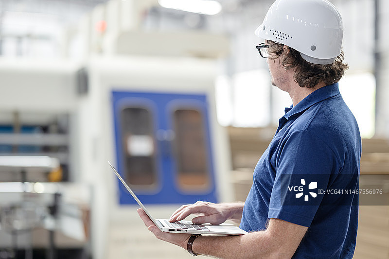 瓦楞纸包装行业的生产和制造管理：生产经理或企业主使用笔记本电脑，同时查看瓦楞纸箱印刷机，以推动和改进业务流程的后视图图片素材