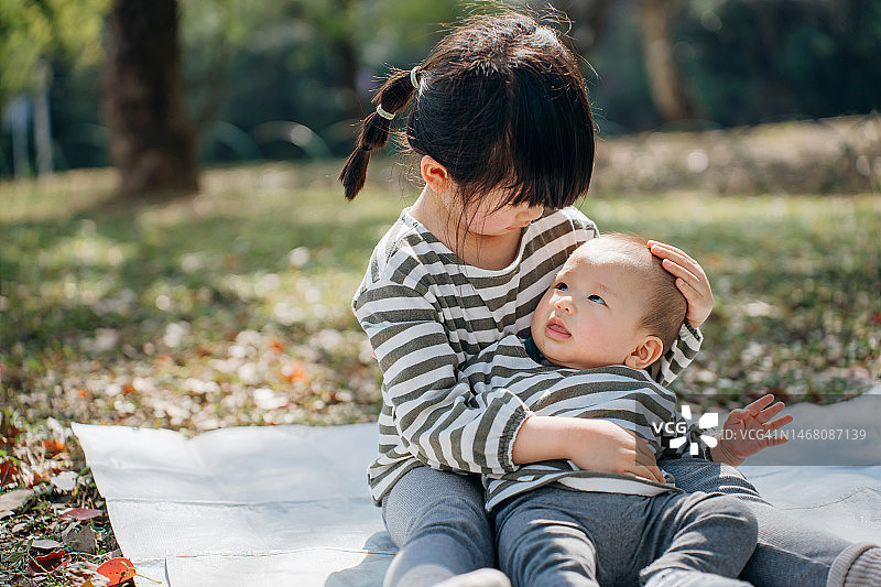 一位亚洲女孩在野餐垫上拥抱她的妹妹，在自然公园里玩耍。快乐的姐妹俩在春光明媚的季节野餐放松，手足情深图片素材