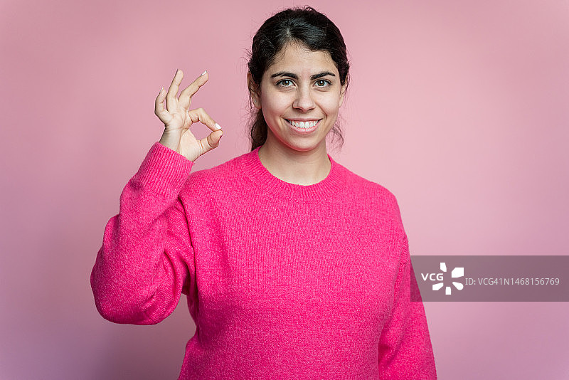 身穿毛衣，在粉色背景前做OK手势的女人图片素材