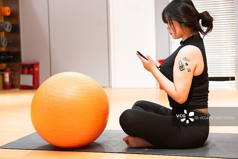 在健身房垫子上锻炼后使用手机的年轻漂亮亚洲女人图片素材