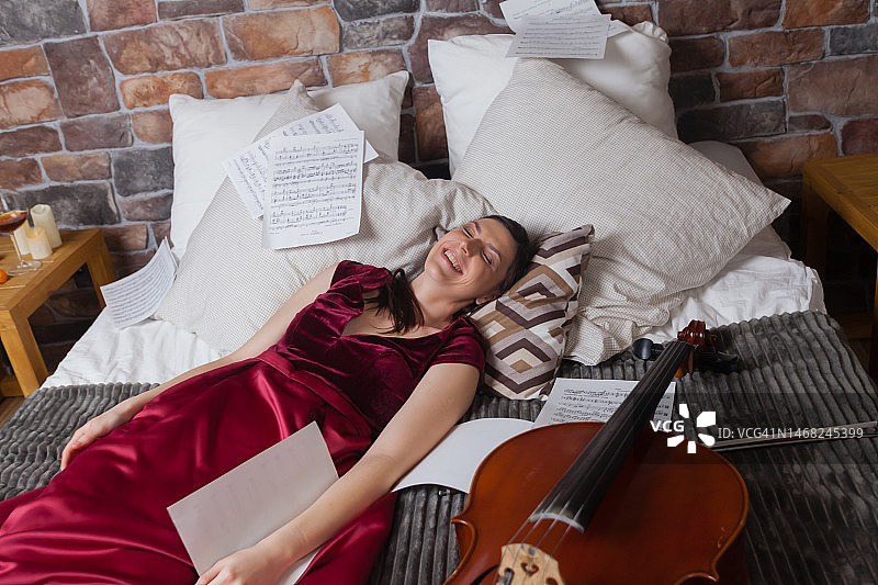 一位女士穿着红色连衣裙躺在床上，看着乐谱，旁边有一把大提琴，背景是砖墙图片素材