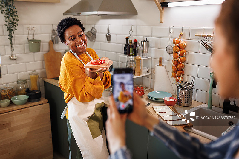 朋友拍摄年轻漂亮的女子，她戴着围裙，拿着放有樱桃番茄的砧板，并向镜头展示图片素材
