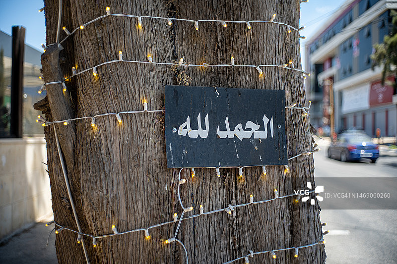 装饰着灯串的街道上，树上写着“如果上帝愿意”的黑色木牌图片素材