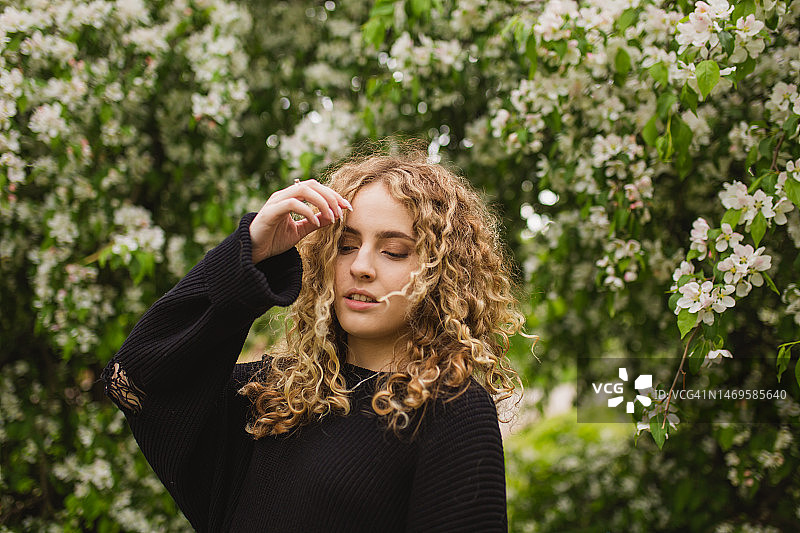 在开花的树背景下，身穿黑色毛衣的金发卷发女孩。花园。白色花朵图片素材