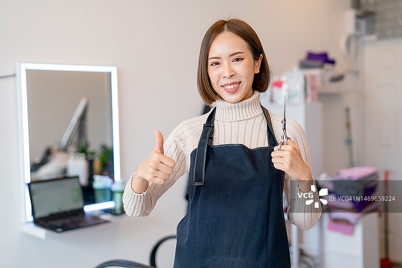 亚裔理发女工肖像，拿着剪刀自信地站着，看着镜头也高兴地竖起大拇指图片素材