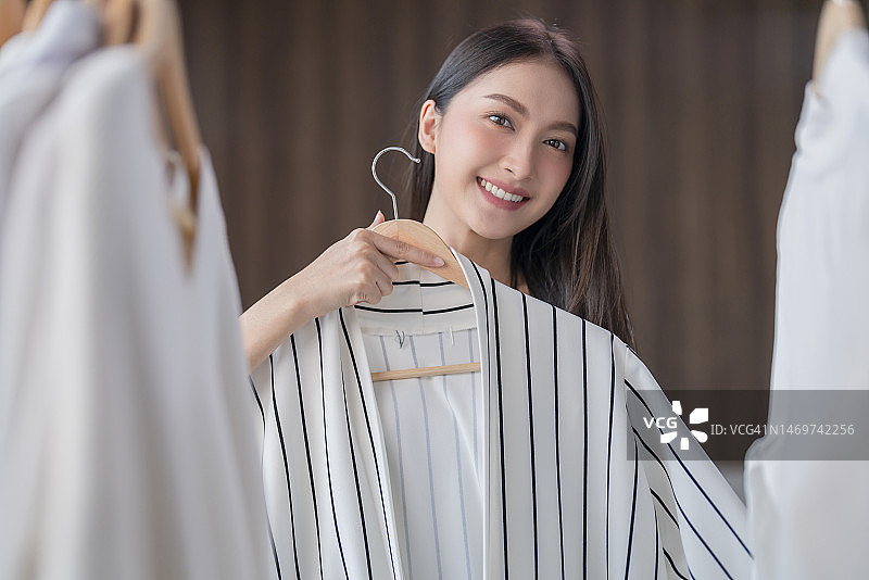 有魅力的亚洲成年年轻女子在家中选择并整理衣架上干净的新衣服，亚洲家庭主妇用手移动衣架上的衣服图片素材