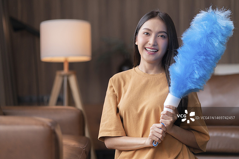 年轻的亚洲家庭主妇在客厅用鸡毛掸子清洁沙发图片素材
