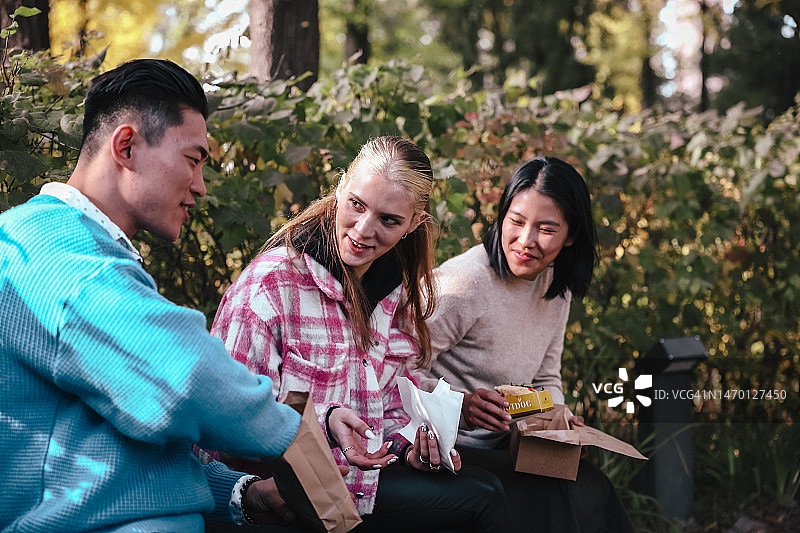 快乐的亚洲和欧洲朋友在首尔纯素街头美食节的餐饮摊位城市公园的户外长椅上聊天、吃纯素食品图片素材