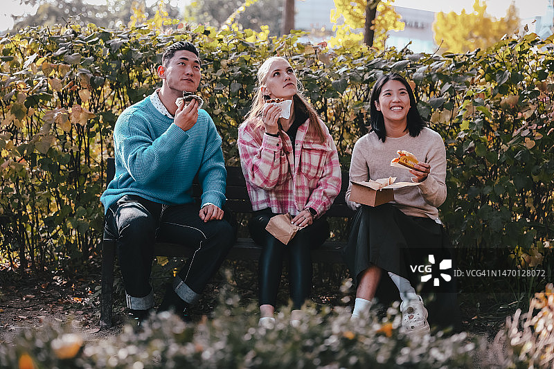 快乐的亚洲和欧洲朋友在首尔纯素街头美食节的餐饮摊位城市公园的户外长椅上聊天、吃纯素食品图片素材