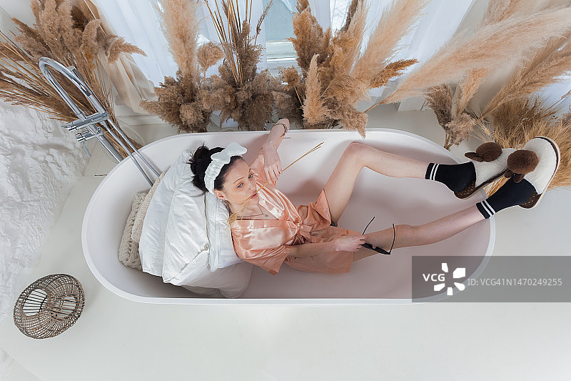 迷人的苗条女孩穿着浴袍，头上戴着蝴蝶结，穿着袜子和拖鞋，躺在浴缸里。图片素材