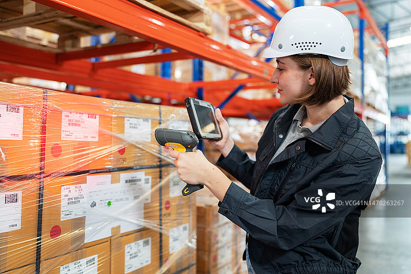 应用RFID标签助力便捷交付，可持续发展业务。戴安全帽的白人仓库工人使用条形码读取器扫描箱子上的二维码，站在配送仓库中，看着平板电脑图片素材